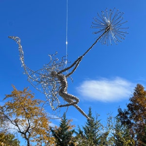 May include: A whimsical, silver wire sculpture of a figure reaching towards a spherical, spiky form. The sculpture is suspended against a bright blue sky, with trees in the background. The artwork evokes a sense of movement and grace.