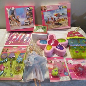 Puede incluir: Una colección de artículos de la marca Barbie que incluye dos cajas de rompecabezas rectangulares, una muñeca con vestido azul, un juguete electrónico rosa con forma de flor, un organizador de plástico verde y varios accesorios en envases tipo blíster.