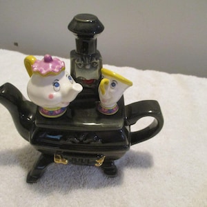 Può includere: Una teiera in ceramica nera a forma di stufa vintage, con sopra una piccola teiera bianca con coperchio rosa e una piccola tazza bianca con bordo giallo. Sia la teiera che la tazza presentano tratti del viso dipinti e dettagli colorati.