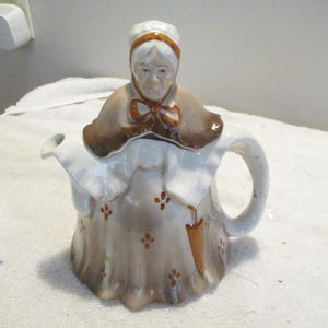 Puede incluir: Una tetera de cerámica vintage con forma de anciana. La tetera presenta una combinación de colores blanco y marrón con detalles florales y un asa. La cabeza de la mujer forma la tapa y el pico está en el lado izquierdo.