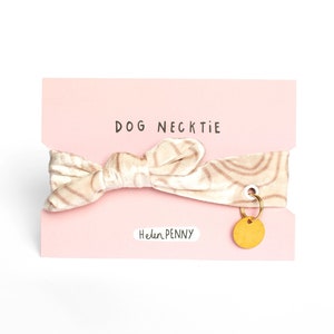 May include: A dog necktie on a pink card. The necktie is beige with a swirl pattern and a bow shape. A small gold-colored charm hangs from the card. The text "DOG NECKTIE" is printed at the top, and "Helen Penny" is printed at the bottom.