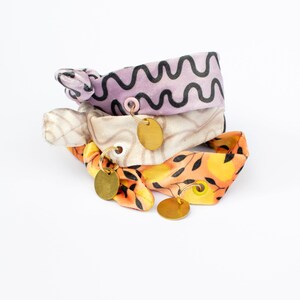 May include: Three fabric hair ties in a stack. The top tie is lavender with a black wavy pattern. The middle tie is beige with a light brown pattern. The bottom tie is orange with a black leaf pattern. Each tie has a gold-colored charm.