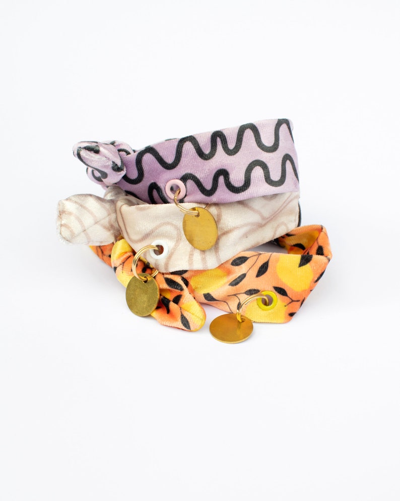 May include: Three fabric hair ties in a stack. The top tie is lavender with a black wavy pattern. The middle tie is beige with a light brown pattern. The bottom tie is orange with a black leaf pattern. Each tie has a gold-colored charm.