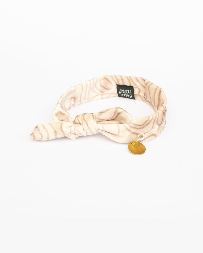 May include: A cream-colored headband with a brown, wood grain-like pattern. The headband is tied in a knot and features a small, gold-colored circular charm. The brand name "Helen Penny" is printed on a black tag.