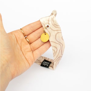 May include: A beige headband with a brown, topographic-like pattern. A small, gold-colored circular tag hangs from the headband. The tag is attached with a gold-colored ring. The brand name "Helen Penny" is printed on a black tag.