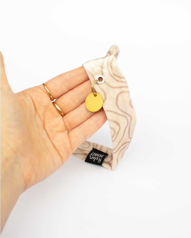 May include: A beige headband with a brown, topographic-like pattern. A small, gold-colored circular tag hangs from the headband. The tag is attached with a gold-colored ring. The brand name "Helen Penny" is printed on a black tag.