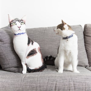 May include: Two cats, one a plush toy and the other a real cat, sit on a gray couch. The plush cat is white with brown spots and has a blue collar. The real cat is white with brown spots and has a blue collar with a bell.