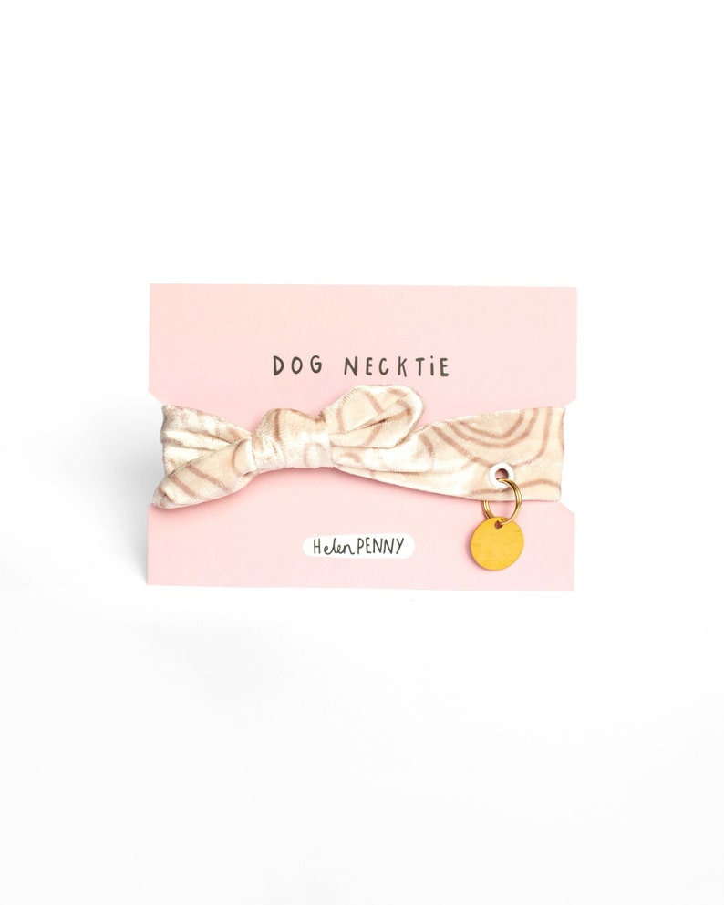 May include: A dog necktie on a pink card. The necktie is beige with a swirl pattern and a bow shape. A small gold-colored charm hangs from the card. The text "DOG NECKTIE" is printed at the top, and "Helen Penny" is printed at the bottom.