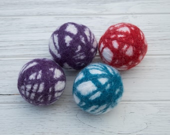 May include: Four felted wool dryer balls in shades of purple, red, and teal, each with a white patterned design. The balls are arranged on a white wooden surface, offering a natural and eco-friendly laundry solution.