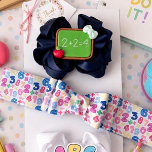 May include: Three hair bows on a white card. The top bow is navy blue with a green chalkboard charm displaying 2+2=4 and a red apple. The middle bow features a white ribbon with colourful math symbols and numbers. The bottom bow is white with an ABC charm.