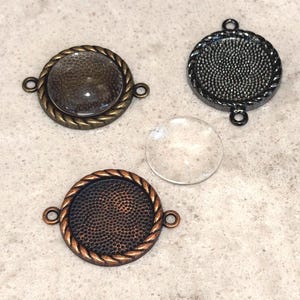 May include: Three round metal jewelry bezels in bronze, black, and copper tones, each with a textured interior and a rope-like border. A clear glass cabochon sits nearby. The bezels have small loops for attaching to other components.