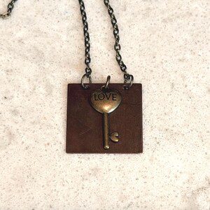 May include: A bronze-toned necklace featuring a square pendant with a key charm. The key has a heart-shaped top with the word "LOVE" and a heart-shaped detail at the bottom. The chain is a dark, linked style.