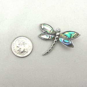 May include: A silver dragonfly brooch with iridescent abalone shell wings and a small clear gemstone. The brooch is next to a U.S. dime coin for size comparison. The dragonfly's wings display shades of green, blue, and silver.