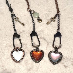 May include: Three heart-shaped pendant necklaces with antique bronze chains. Each pendant has a different colored heart: white, orange, and pink. Each necklace includes a small key charm and a metal clasp.