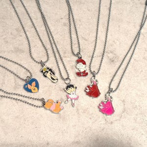 May include: A collection of silver-toned necklaces with various cartoon character pendants. The pendants include a blue heart with a bunny, a car, a Smurf, a ballerina, and princesses in red and pink dresses. The necklaces are on a light-colored surface.