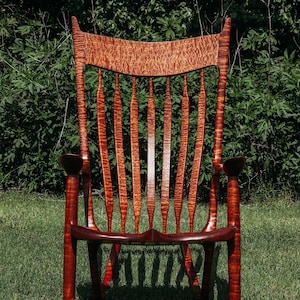 May include: A wooden rocking chair with a high back and intricate details. The chair is made of a reddish-brown wood with a natural grain pattern. The chair is sitting on a grassy lawn.
