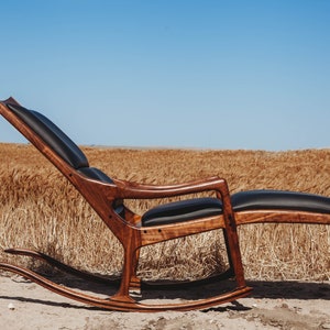 May include: A wooden rocking chair with a black leather seat and footrest. The chair is in a field of tall grass.