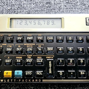 Könnte beinhalten: Ein schwarz-goldener HP 12C Finanzrechner mit einem digitalen Display, das die Zahl 123.456.789 anzeigt. Der Rechner verfügt über eine Vielzahl von Tasten, darunter AMORT, INT, NPV, RND, IRR, CHS, DATE, BEG, END, MEM, PRICE, YTM, SL, SOYD, DB, EEX, ADYS, D.MY, M.DY, P/R, Σ, PRGM, CLEAR, FIN, REG, PREFIX, R/S, SST, PSE, BST, R, GTO, xy, x=0, xzy, CLX, ON, f, g, STO, RCL, LSTX, ENTER, 1, 2, 3, -, +, 0, ., Σ+, und +.