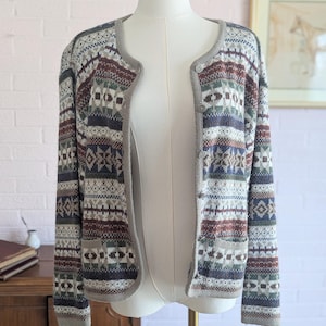 May include: A patterned cardigan in shades of gray, burgundy, green, and blue, displayed on a mannequin. The open-front sweater features a round neckline and a button closure. The design includes geometric and stylized floral motifs.