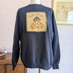 May include: Navy blue sweatshirt with a "TED E. BEAR" patch featuring a teddy bear wearing a hat. The patch is rectangular with a frayed edge. The teddy bear is tan with pink accents.