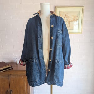 May include: A dark blue denim jacket with a brown corduroy collar and large front pockets. The cuffs are rolled up to reveal a red and black plaid lining. The jacket has large wooden buttons and is displayed on a mannequin.