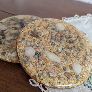 May include: Two round coasters with a beach theme. Each coaster is embedded with sand, small seashells, and pebbles, creating a textured surface. The coasters have a clear resin coating and a brown border. The coasters are on a wooden surface.