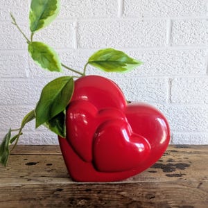 May include: A glossy red heart-shaped vase with two smaller hearts embossed on the front. Green leafy plant stems emerge from the top. The vase sits on a wooden surface against a white brick wall.