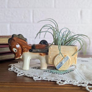 May include: A ceramic donkey planter with a small wooden cart. The donkey is brown and white with a black saddle. The cart is beige and holds a green plant. The planter is on a wooden surface.