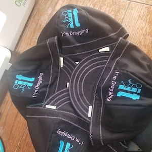 May include: Four black welding caps with a blue embroidered design of a stick figure dragging a toilet. The text "I'm dragging it" is embroidered on each cap.