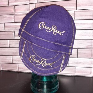 Puede incluir: Una gorra de ciclismo morada con el texto Crown Royal bordado en dorado tanto en la corona como en la visera. La gorra presenta costuras en zigzag de color dorado visibles a lo largo de las uniones, creando un contraste con la tela morada.