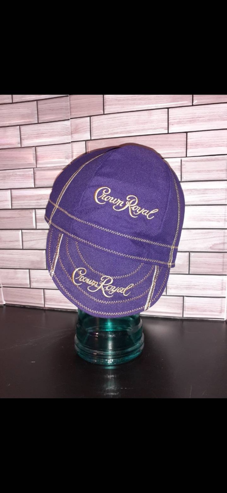 Puede incluir: Una gorra de ciclismo morada con el texto Crown Royal bordado en dorado tanto en la corona como en la visera. La gorra presenta costuras en zigzag de color dorado visibles a lo largo de las uniones, creando un contraste con la tela morada.