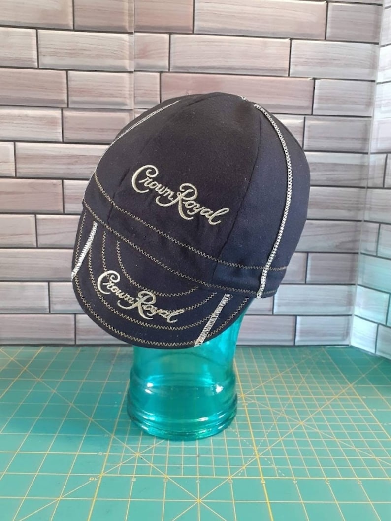 May include: Black Crown Royal branded welding cap with gold embroidery. The cap has a brim and a seam down the center of the crown.