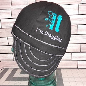 May include: Black welding cap with a turquoise blue embroidered design of a stick figure dragging a toilet. The text "I'm Draggling" is embroidered below the design.