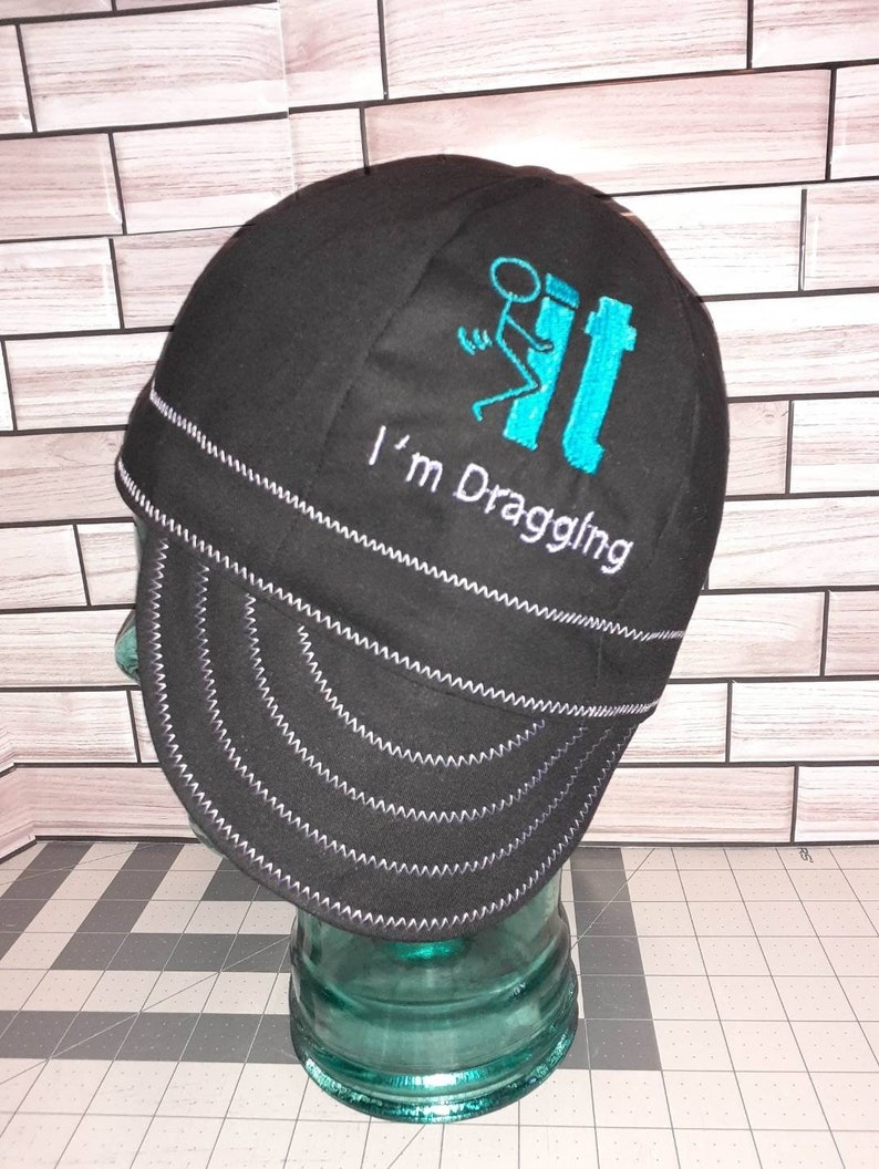 May include: Black welding cap with a turquoise blue embroidered design of a stick figure dragging a toilet. The text "I'm Draggling" is embroidered below the design.