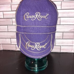 Puede incluir: Una gorra de ciclismo morada con el logotipo de Crown Royal bordado en dorado en el panel frontal y en la visera. La gorra presenta costuras en zigzag de color dorado visibles a lo largo de las uniones, creando un contraste con la tela.