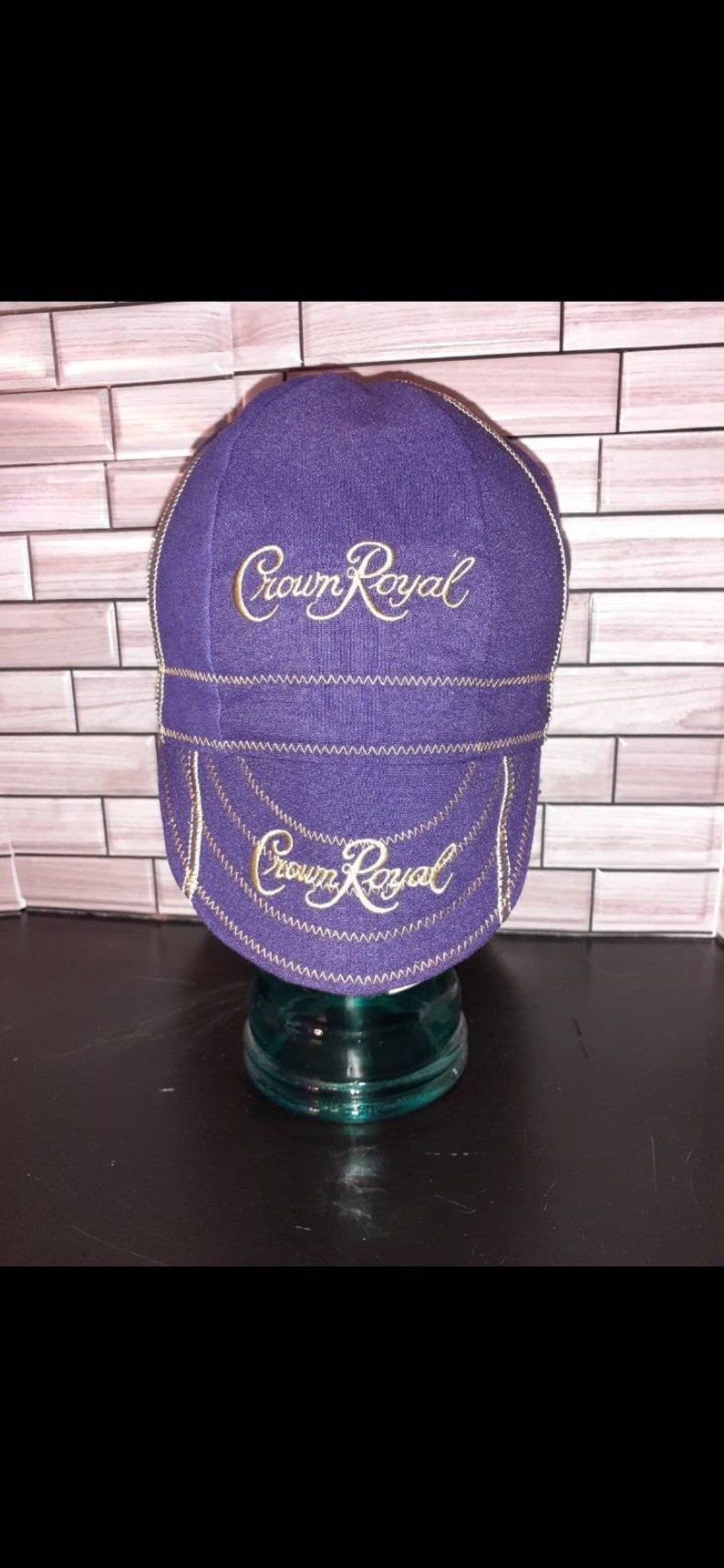 Puede incluir: Una gorra de ciclismo morada con el logotipo de Crown Royal bordado en dorado en el panel frontal y en la visera. La gorra presenta costuras en zigzag de color dorado visibles a lo largo de las uniones, creando un contraste con la tela.