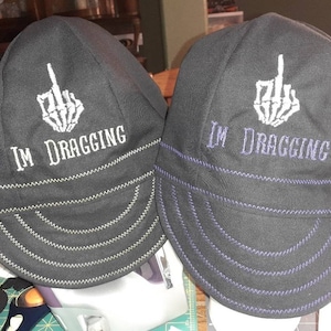 May include: Two black welding caps with a white skeleton middle finger and the text "I'm dragging" embroidered on the front. The cap on the left has green stitching and the cap on the right has purple stitching.