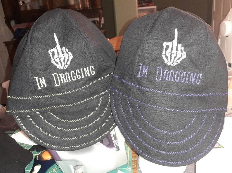 May include: Two black welding caps with a white skeleton middle finger and the text "I'm dragging" embroidered on the front. The cap on the left has green stitching and the cap on the right has purple stitching.
