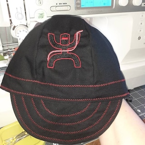 May include: A black and red baseball cap with a red embroidered logo of a stylized figure with a lasso. The cap has a red stitched brim.