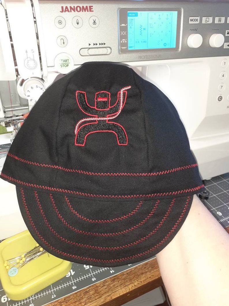 May include: A black and red baseball cap with a red embroidered logo of a stylized figure with a lasso. The cap has a red stitched brim.