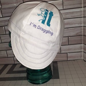 May include: White cotton welding cap with a blue and white stick figure graphic and the text "I'm Dragging" embroidered on the front.