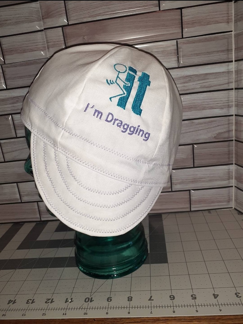 May include: White cotton welding cap with a blue and white stick figure graphic and the text "I'm Dragging" embroidered on the front.