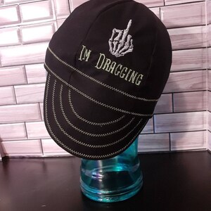 May include: Black welding cap with a white embroidered skeleton hand giving the middle finger and the text "I'M DRAGGING".