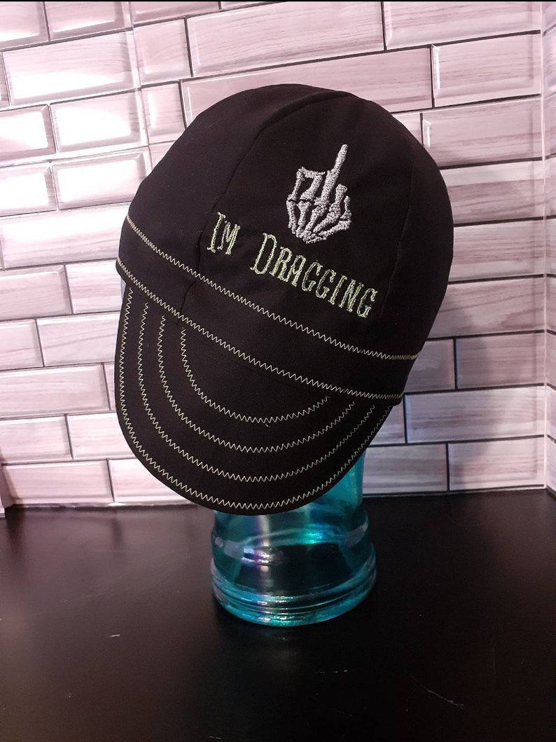 May include: Black welding cap with a white embroidered skeleton hand giving the middle finger and the text "I'M DRAGGING".