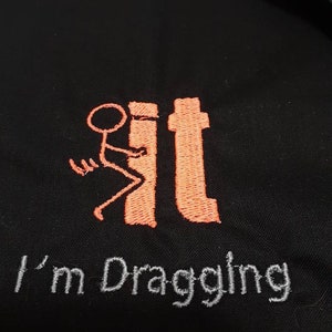 May include: Black fabric with orange embroidery of a stick figure dragging a toilet. The text "I'm Dragging It" is embroidered in gray below the figure.