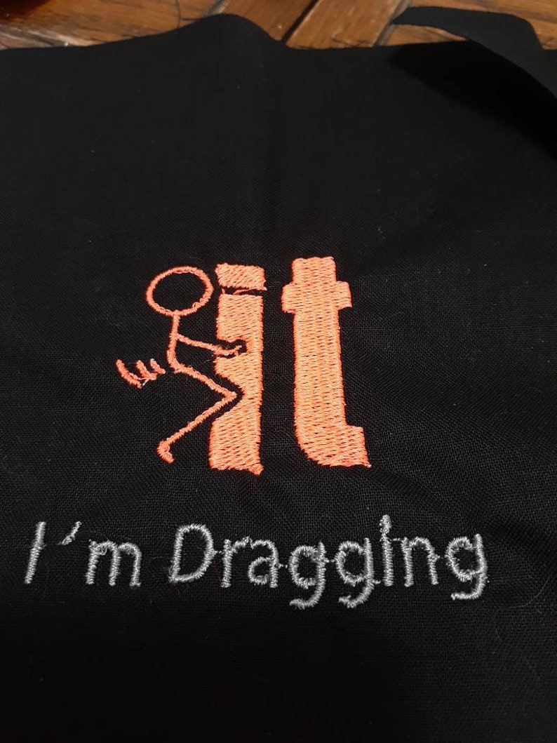 May include: Black fabric with orange embroidery of a stick figure dragging a toilet. The text "I'm Dragging It" is embroidered in gray below the figure.