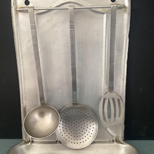 May include: A vintage metal kitchen utensil holder with a ladle, slotted spoon, and skimmer. The holder is a light silver color and has a curved top and a small shelf at the bottom. The utensils have patterned handles.