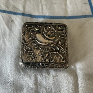 Puede incluir: Una pequeña caja metálica rectangular de color plateado con un intrincado patrón en relieve de hojas de acanto, motivos florales y líneas diagonales ranuradas en la tapa abatible, con un acabado oscuro y oxidado en las zonas hundidas.