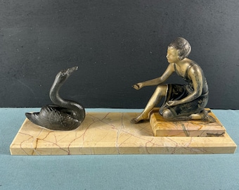 May include: A bronze sculpture of a woman in a black and gold dress sitting on a marble base. A bronze swan sculpture is on the left side of the base.