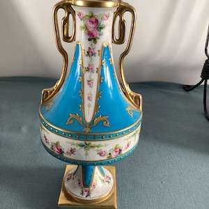 May include: A decorative porcelain vase with a square base and two gold handles. The vase features a vibrant turquoise body, white accents, and delicate pink rose floral patterns. Gold trim and accents complete the design.